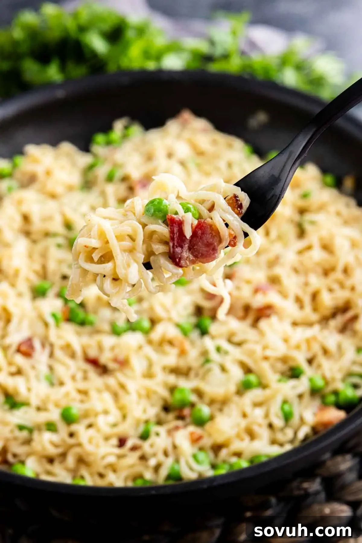 Effortless Ramen Carbonara 5 A fork holds a serving of Easy Ramen Carbonara with peas and bacon over a pan filled with the same noodle mixture.
