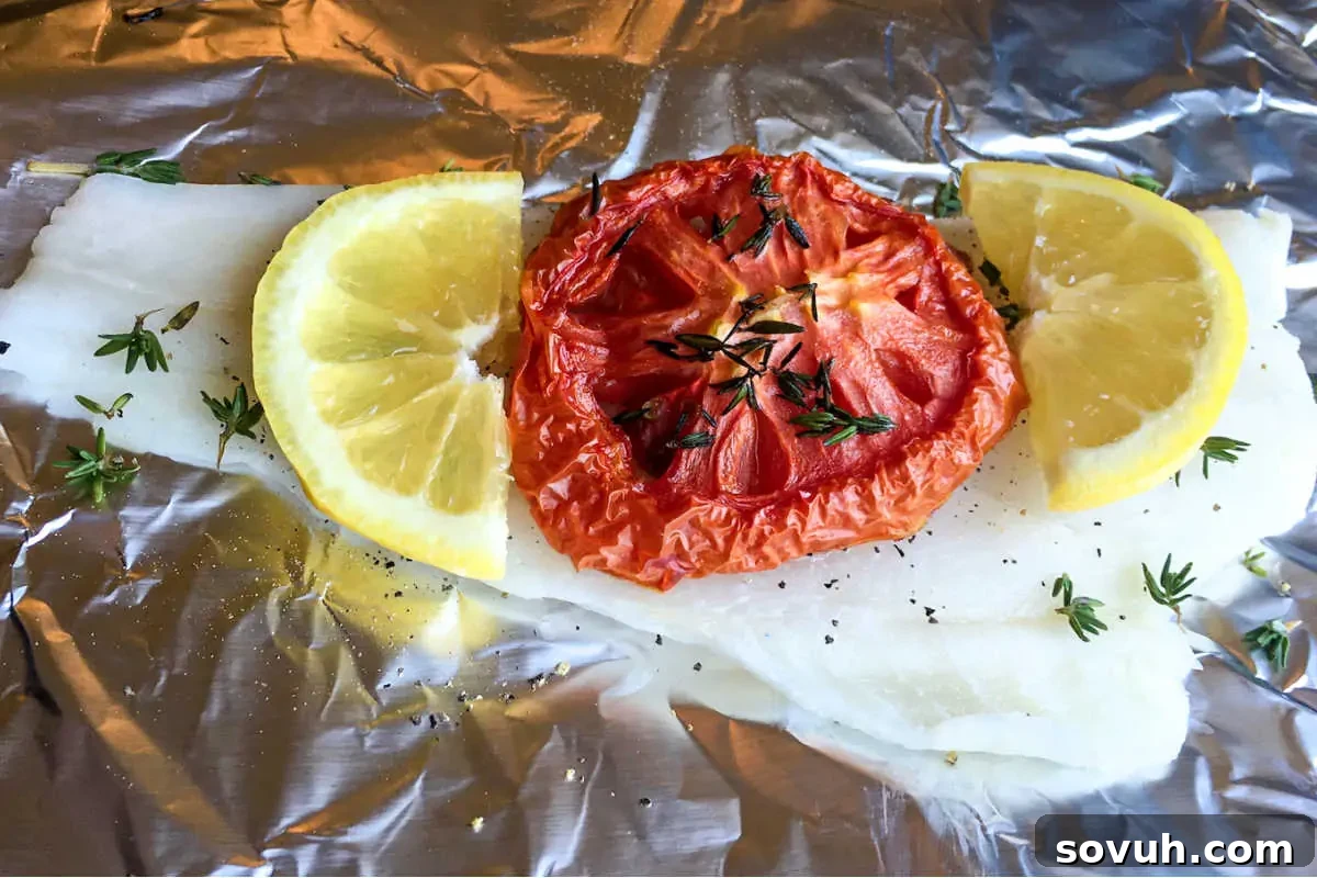 A piece of white fish fillet on foil topped with a roasted tomato slice, flanked by two lemon wedges, and sprinkled with fresh thyme and black pepper.