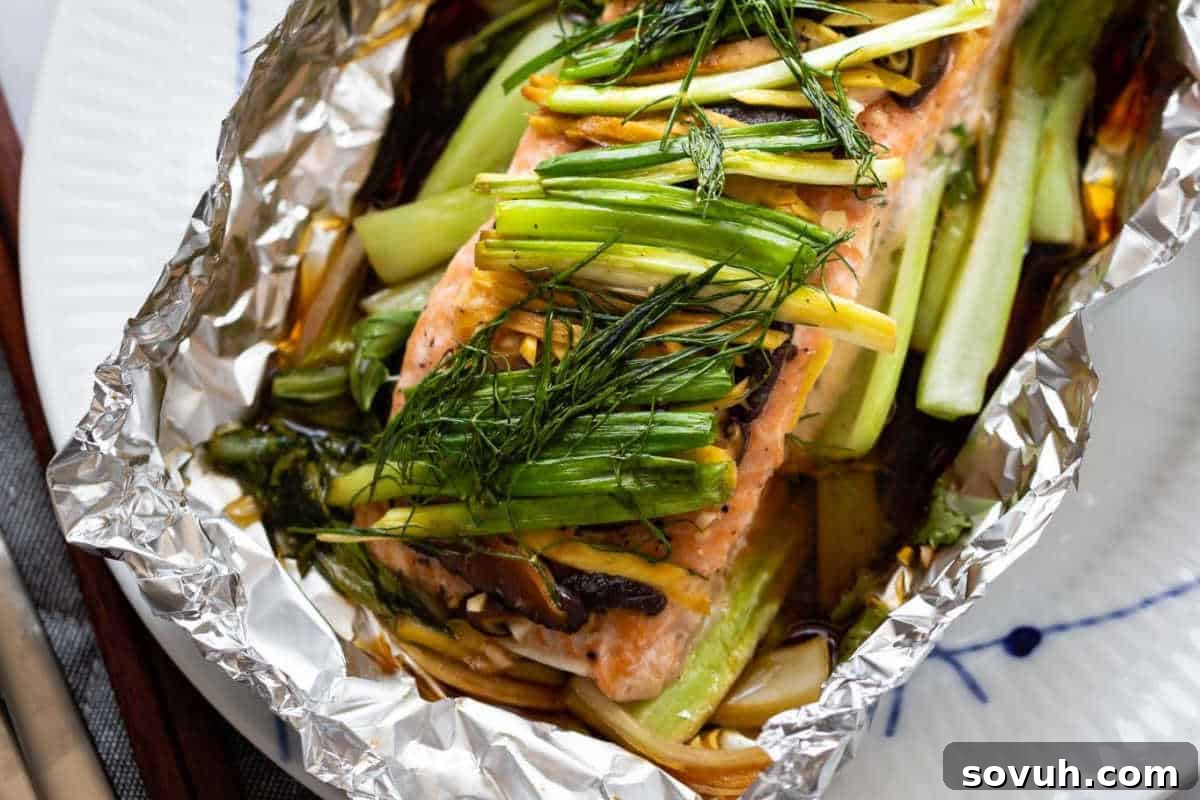 A baked salmon fillet is topped with green onions and dill, served in aluminum foil on a white plate.