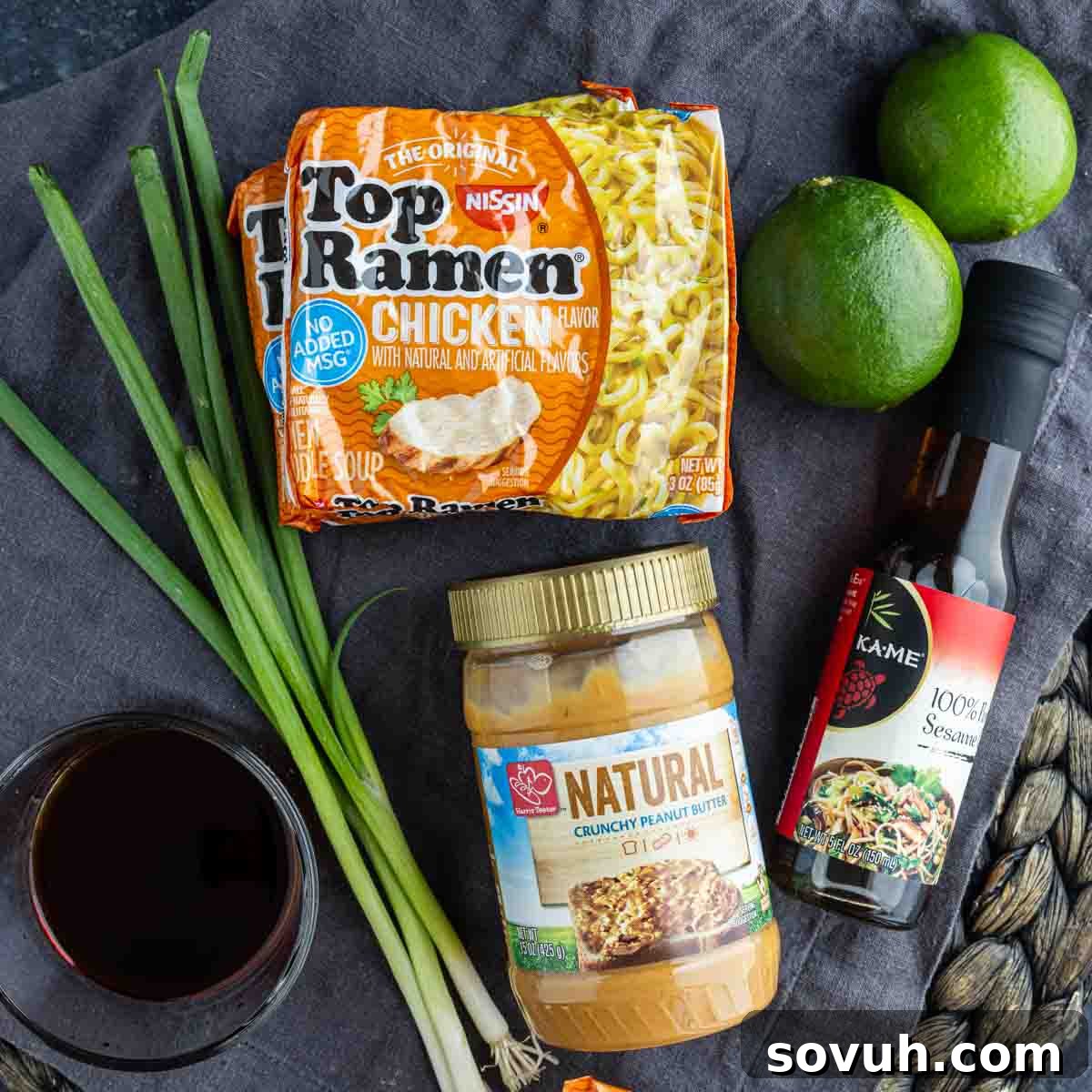 Two packs of chicken-flavored Top Ramen, two limes, green onions, crunchy peanut butter, sesame oil, and a dish of dark liquid set the stage for an easy ramen recipe—perfect for making delicious peanut butter ramen at home.