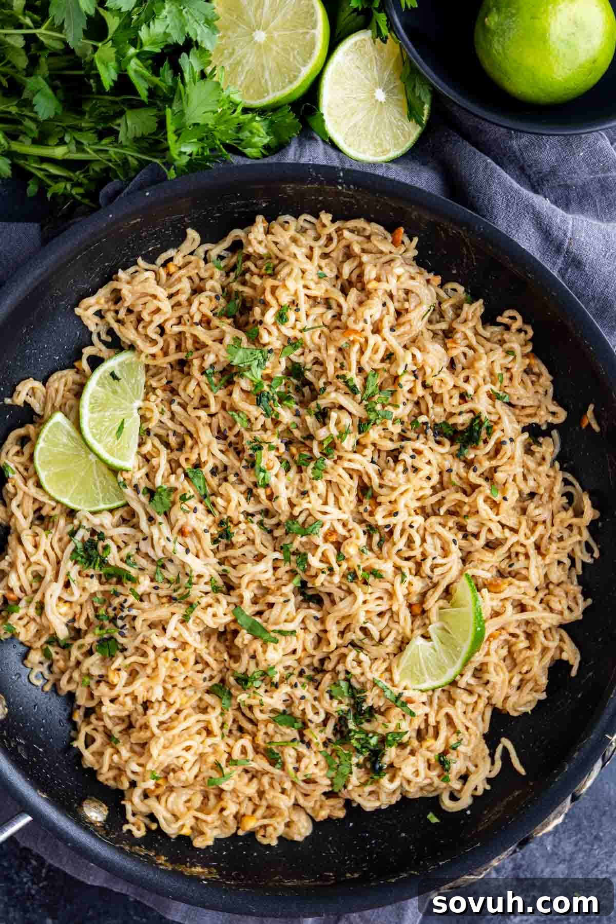 A skillet filled with peanut ramen noodles garnished with chopped herbs and lime wedges, with limes and fresh herbs on the side—an easy ramen recipe for a quick, flavorful meal.