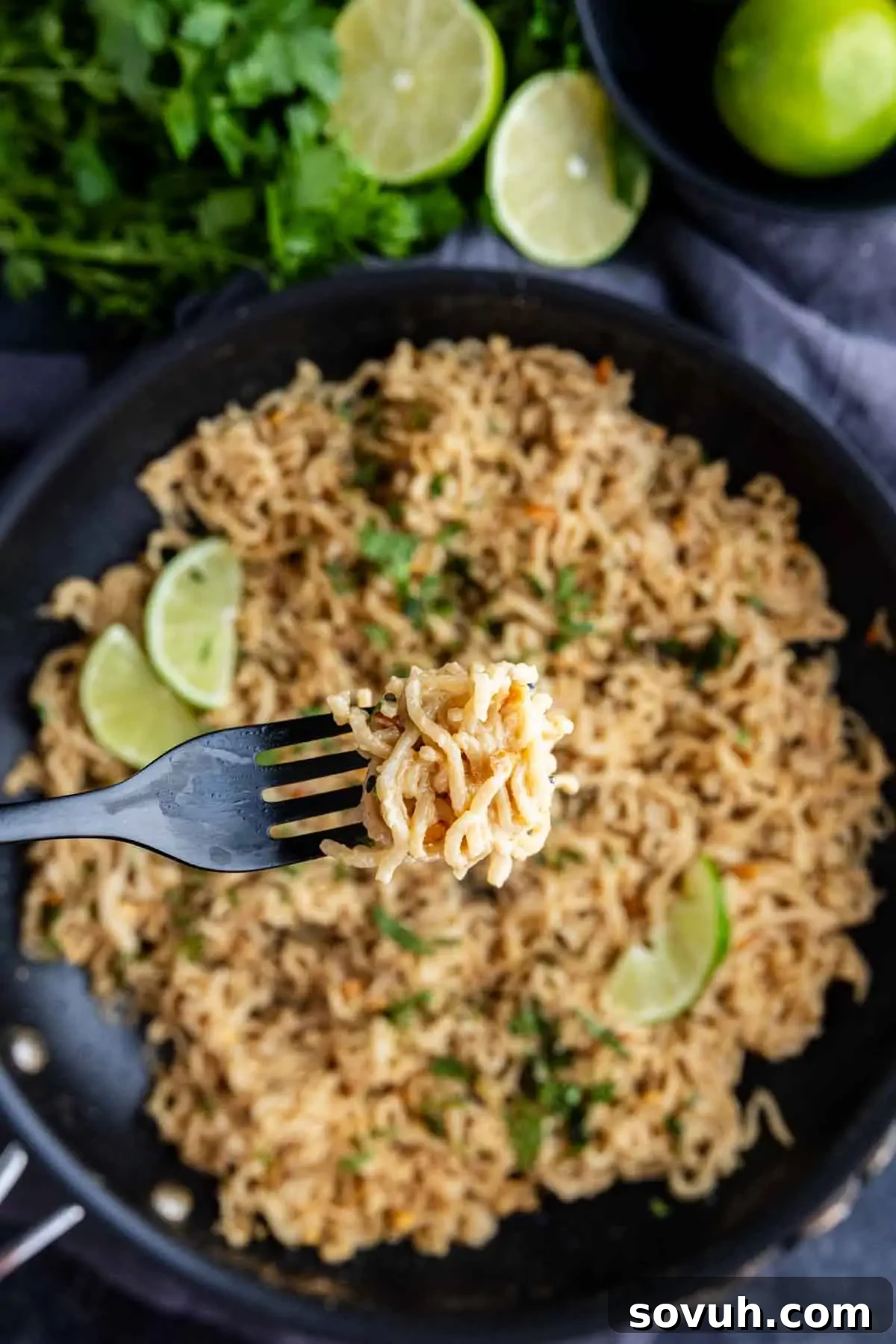 A fork holds a bite of cooked peanut ramen noodles above a pan filled with noodles and lime wedges, with fresh herbs and limes in the background.