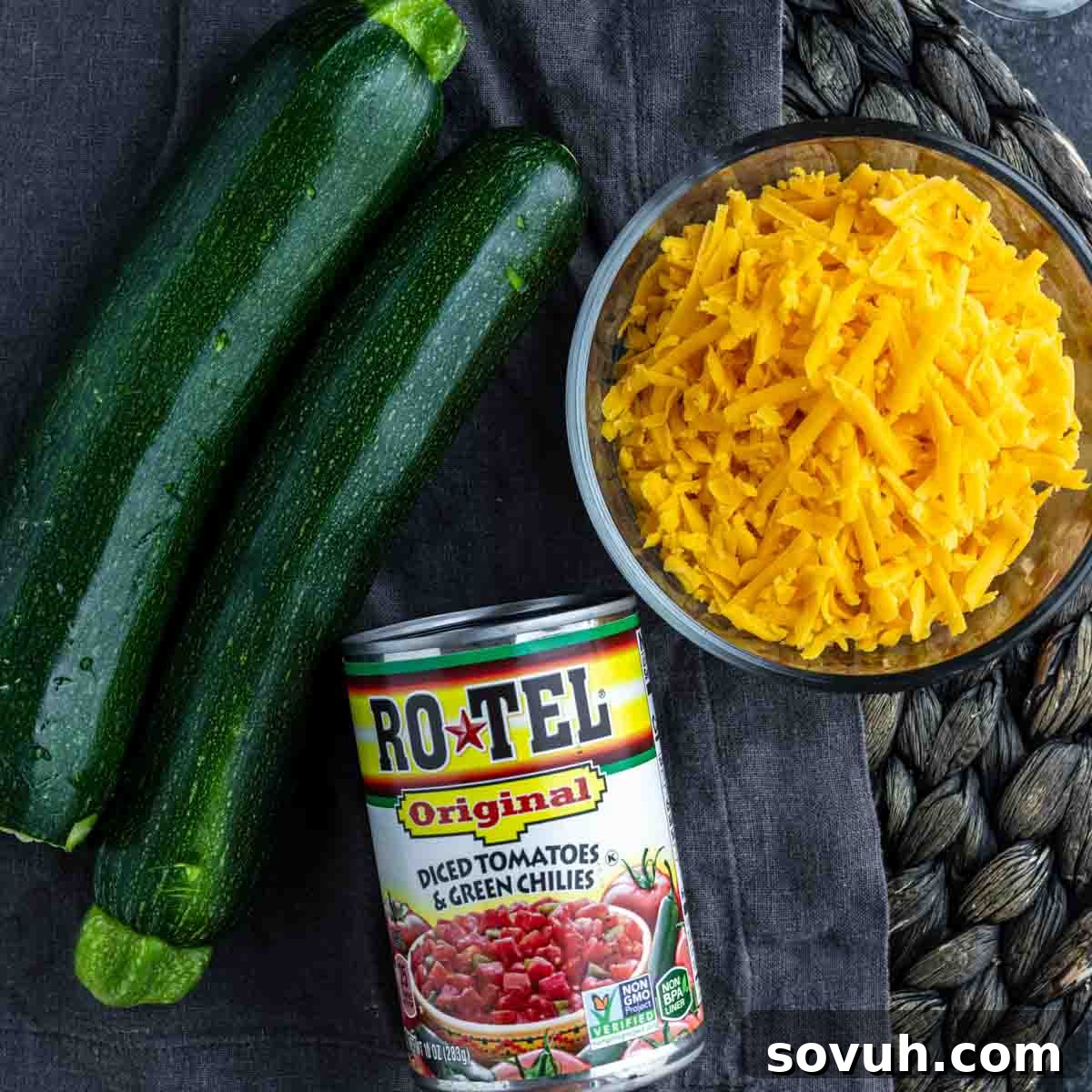 Two whole zucchinis, a can of Rotel diced tomatoes and green chilies, and a bowl of shredded cheddar cheese are ready to create a delicious summer zucchini skillet on a dark cloth surface.