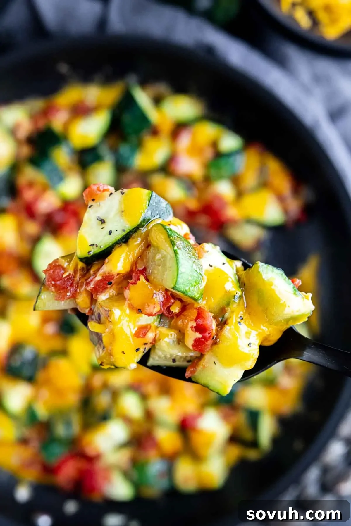 A close-up of a fork holding a bite of zucchini, tomatoes, and melted cheese from a cheesy zucchini skillet, with the pan of this summer zucchini skillet in the background.