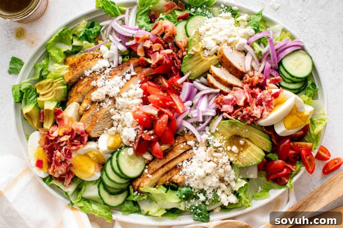 A generously portioned oval white plate holds a vibrant Cobb salad, meticulously arranged with rows of grilled chicken, creamy avocado, perfectly hard-boiled eggs, crispy bacon bits, juicy cherry tomatoes, refreshing cucumber, thinly sliced red onion, and crumbled cheese, creating a visually appealing and satisfying meal.