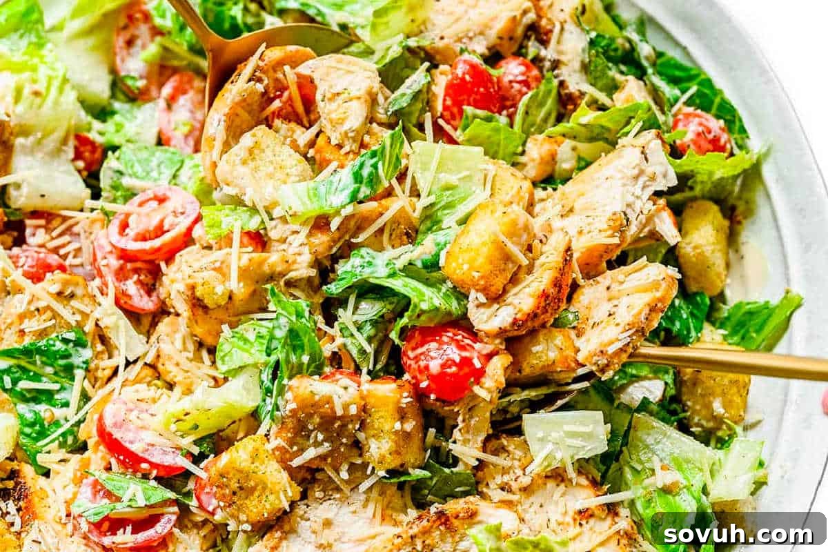 A close-up view of a classic Chicken Caesar salad, featuring crisp romaine lettuce, tender grilled chicken slices, vibrant cherry tomatoes, crunchy golden croutons, and a generous sprinkle of shredded Parmesan cheese, all coated in a rich, creamy Caesar dressing and served in a stylish bowl with elegant gold utensils.