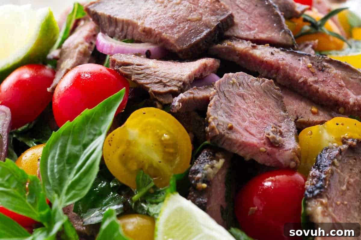 A close-up image of perfectly grilled steak slices, beautifully arranged alongside vibrant cherry tomatoes, thinly sliced red onion, fresh green basil leaves, and bright lime wedges, creating a colorful and enticing presentation.