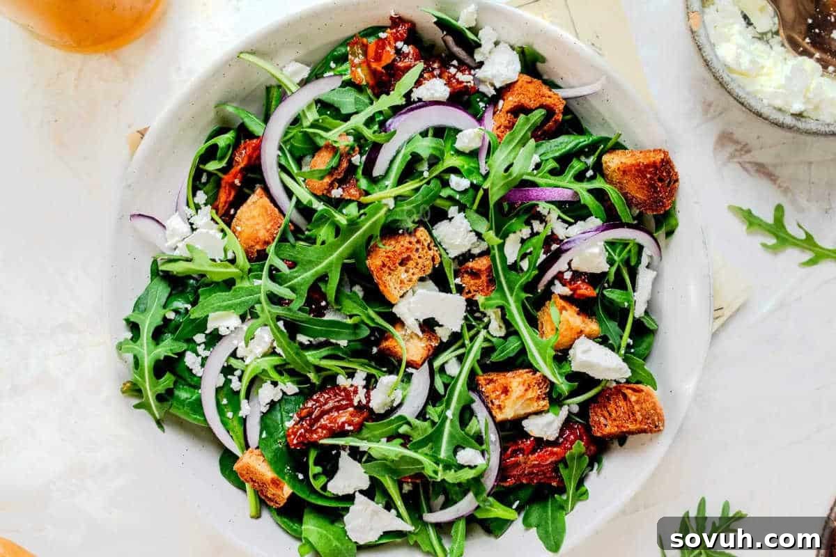 A vibrant bowl of arugula salad, richly adorned with thinly sliced red onion, crumbles of savory feta cheese, succulent sun-dried tomatoes, and crunchy croutons, creating a medley of Mediterranean flavors and textures.