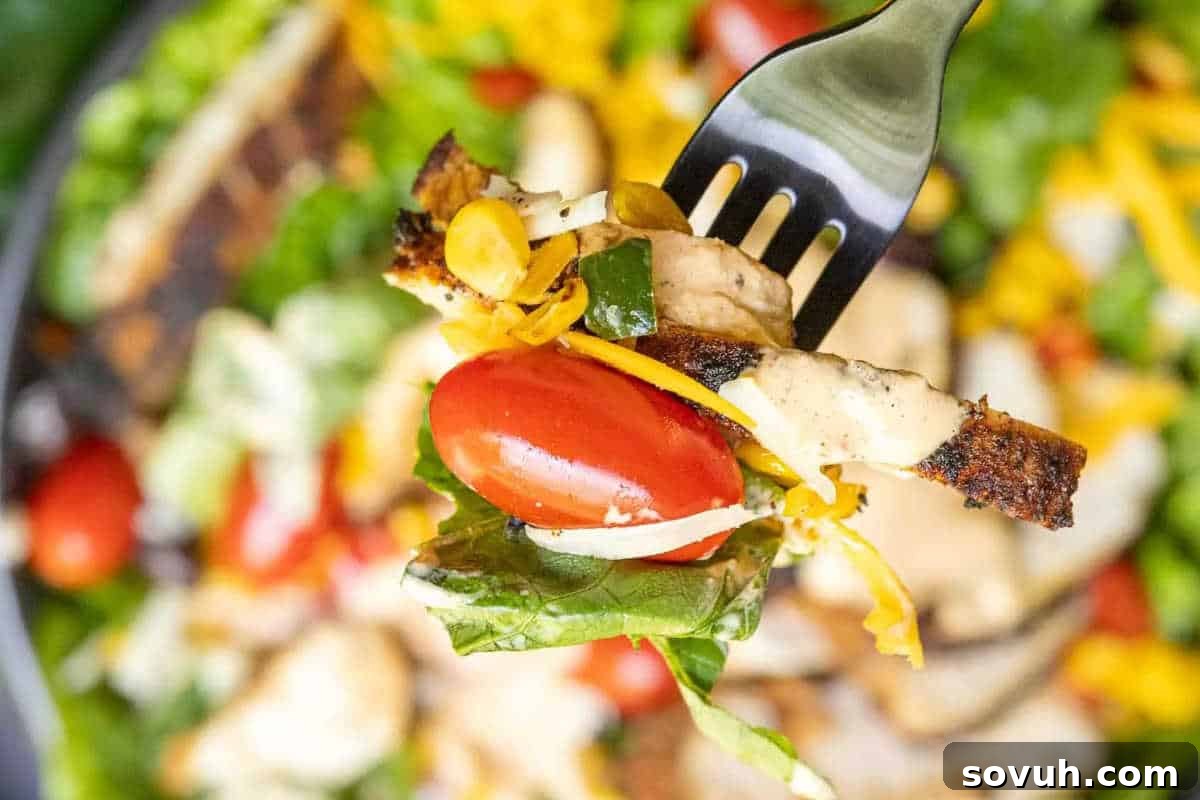 A fork lifts a tantalizing bite of Southwest salad, featuring succulent grilled chicken, fresh crisp lettuce, juicy cherry tomato, sweet corn kernels, and shredded cheese, all generously coated in a creamy, zesty dressing, inviting a taste of its vibrant flavors.
