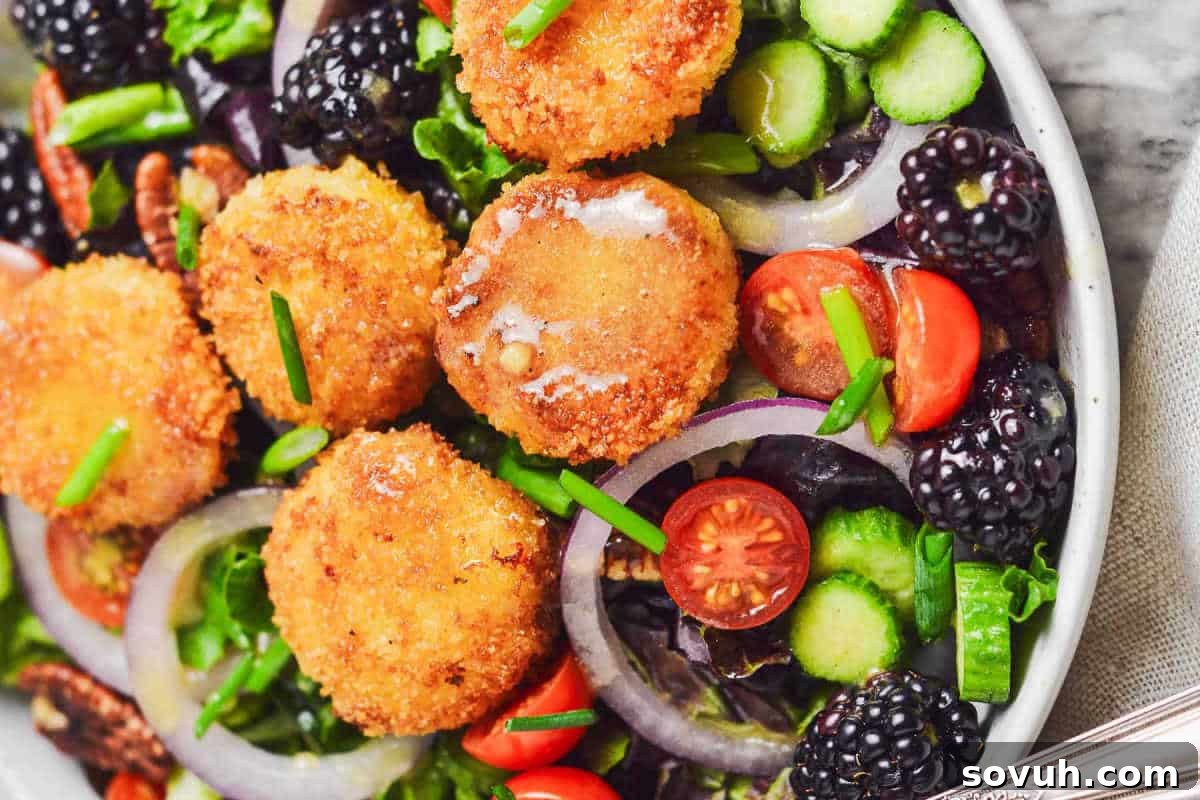 A beautifully composed bowl of salad featuring golden-brown breaded goat cheese rounds, bursting with juicy cherry tomatoes and fresh blackberries, alongside crisp cucumber slices, delicate red onion rings, and a bed of mixed greens, creating a vibrant and textural feast.