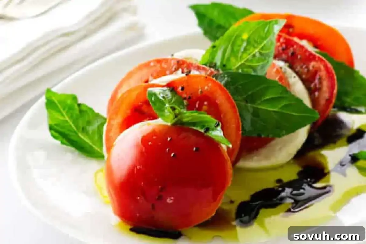 A beautifully arranged plate of classic Caprese salad showcasing thick slices of ripe red tomatoes, creamy fresh mozzarella, and vibrant green basil leaves, lightly drizzled with golden olive oil and dark balsamic vinegar, and garnished with a sprinkle of freshly ground black pepper.