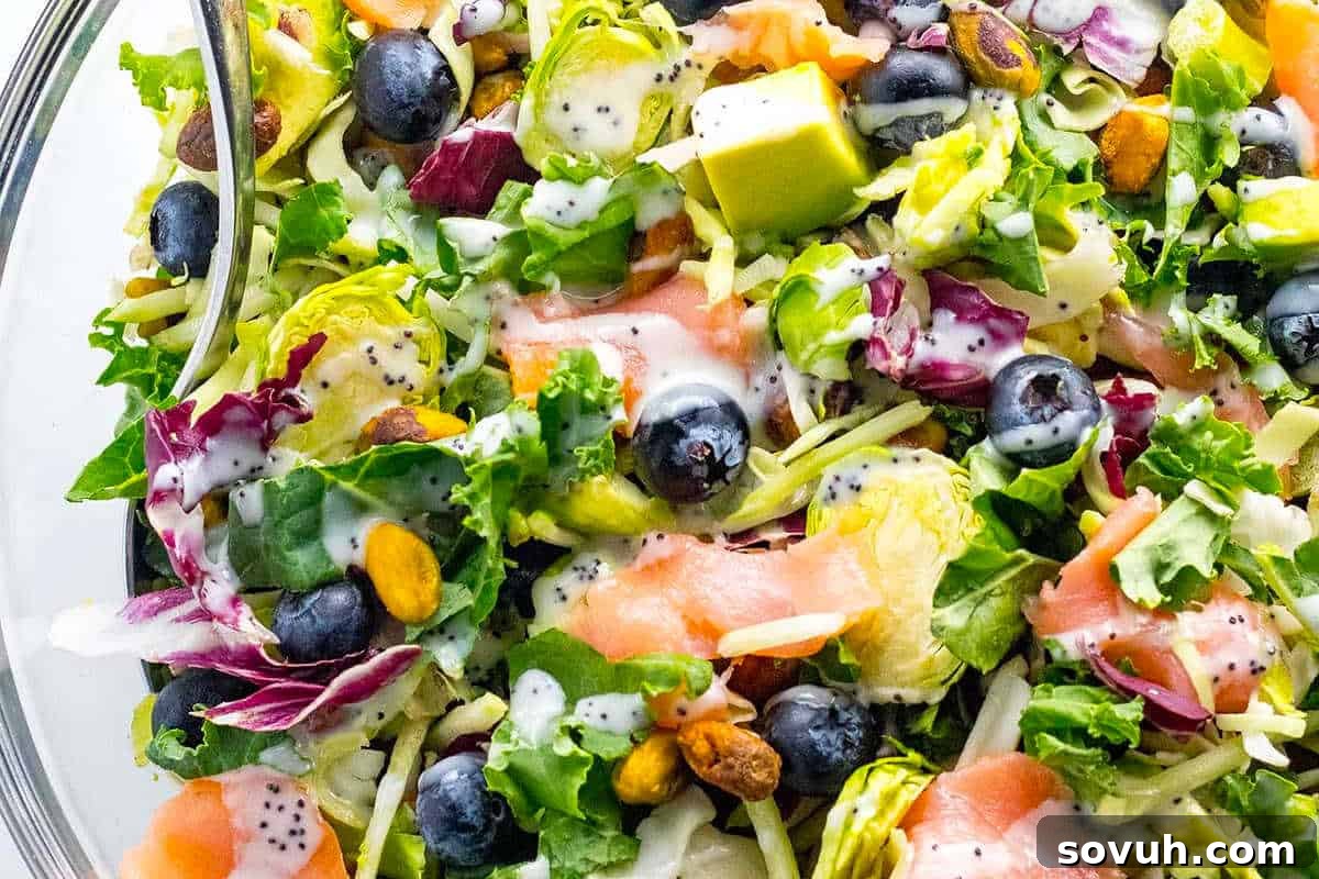 A vibrant mixed salad presented in a clear glass bowl, featuring crisp lettuce, creamy avocado slices, sweet blueberries, crunchy pistachios, succulent smoked salmon pieces, and a generous drizzle of poppy seed dressing, creating a harmonious blend of colors and textures.