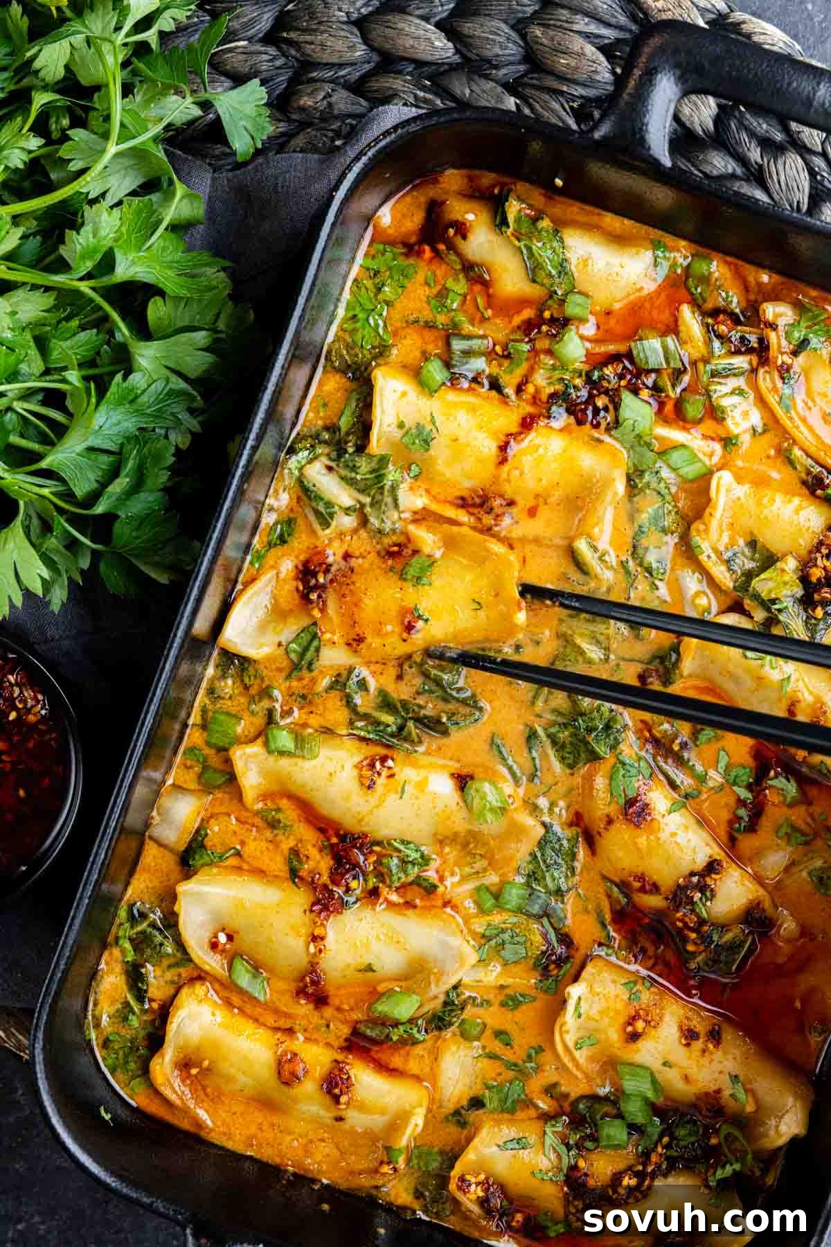 A tray of one pan baked dumplings in a spicy orange broth, garnished with chopped green onions and herbs, with chopsticks picking up one dumpling and fresh parsley on the side.