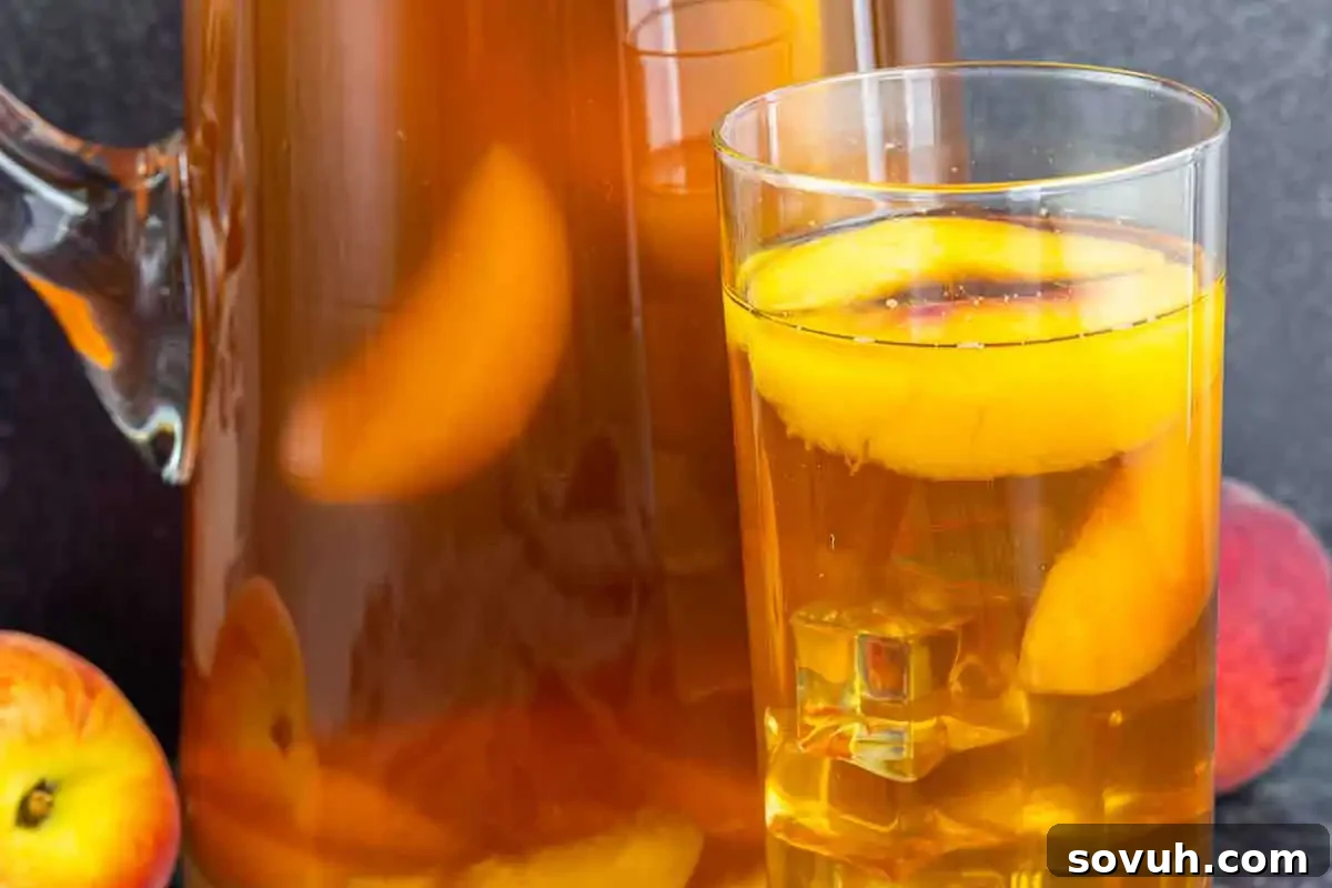 A glass and a pitcher of iced tea with peach slices inside, placed next to whole peaches on a dark surface.