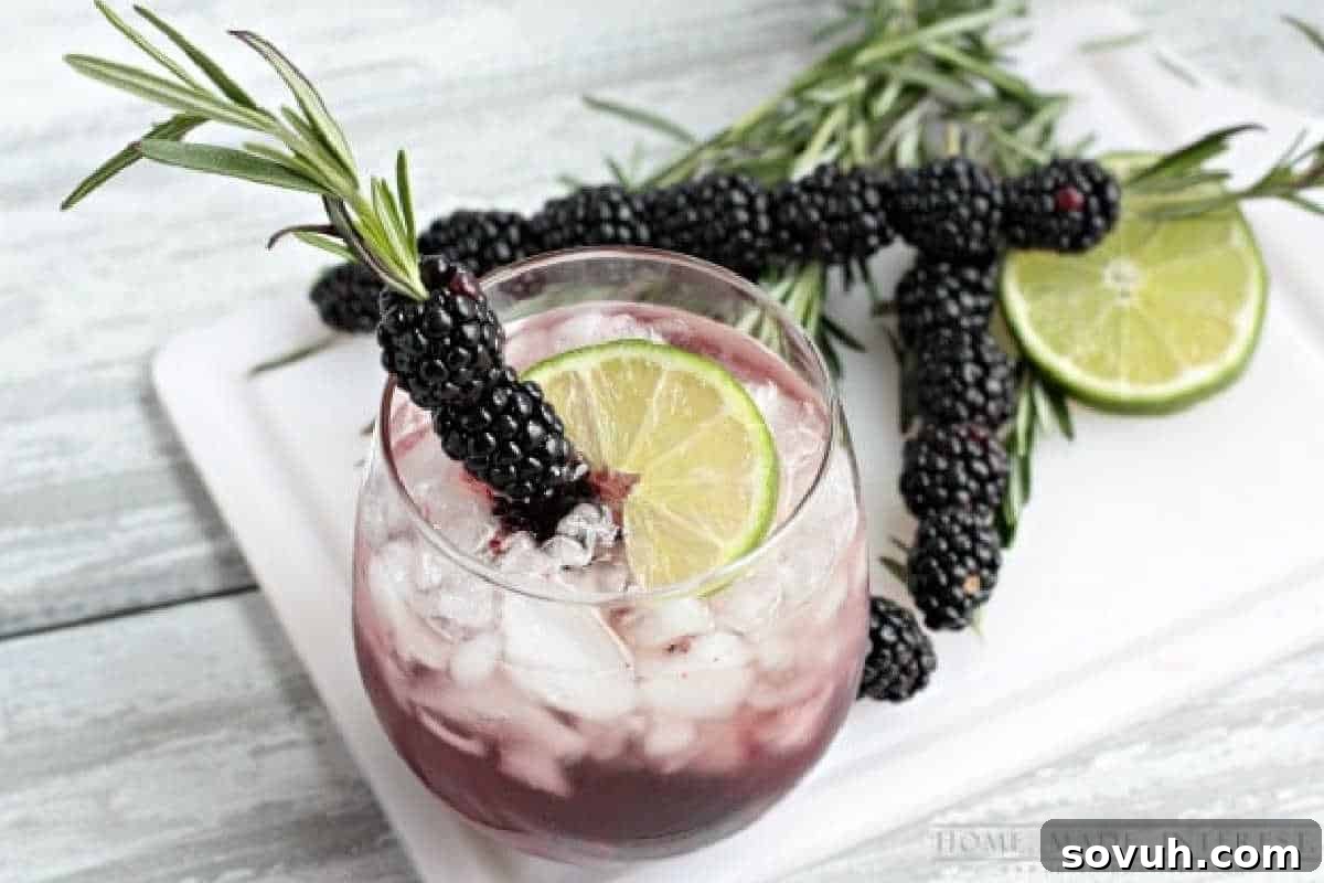 A glass of iced berry cocktail garnished with a lime slice and blackberries on a rosemary sprig, with more blackberries, rosemary, and sliced lime on a white board.