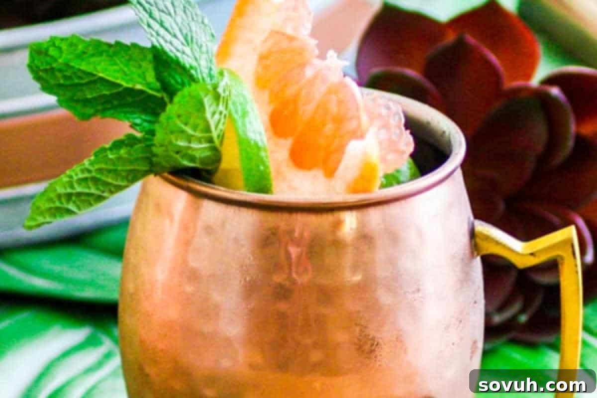 A copper mug filled with a cocktail, garnished with a grapefruit wedge and fresh mint leaves, sits on a green surface with a blurred succulent in the background.