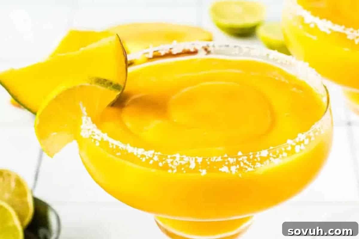 A glass of frozen mango margarita garnished with a lime wedge and a mango slice, with a salted rim. Lime halves and another drink are visible in the background.
