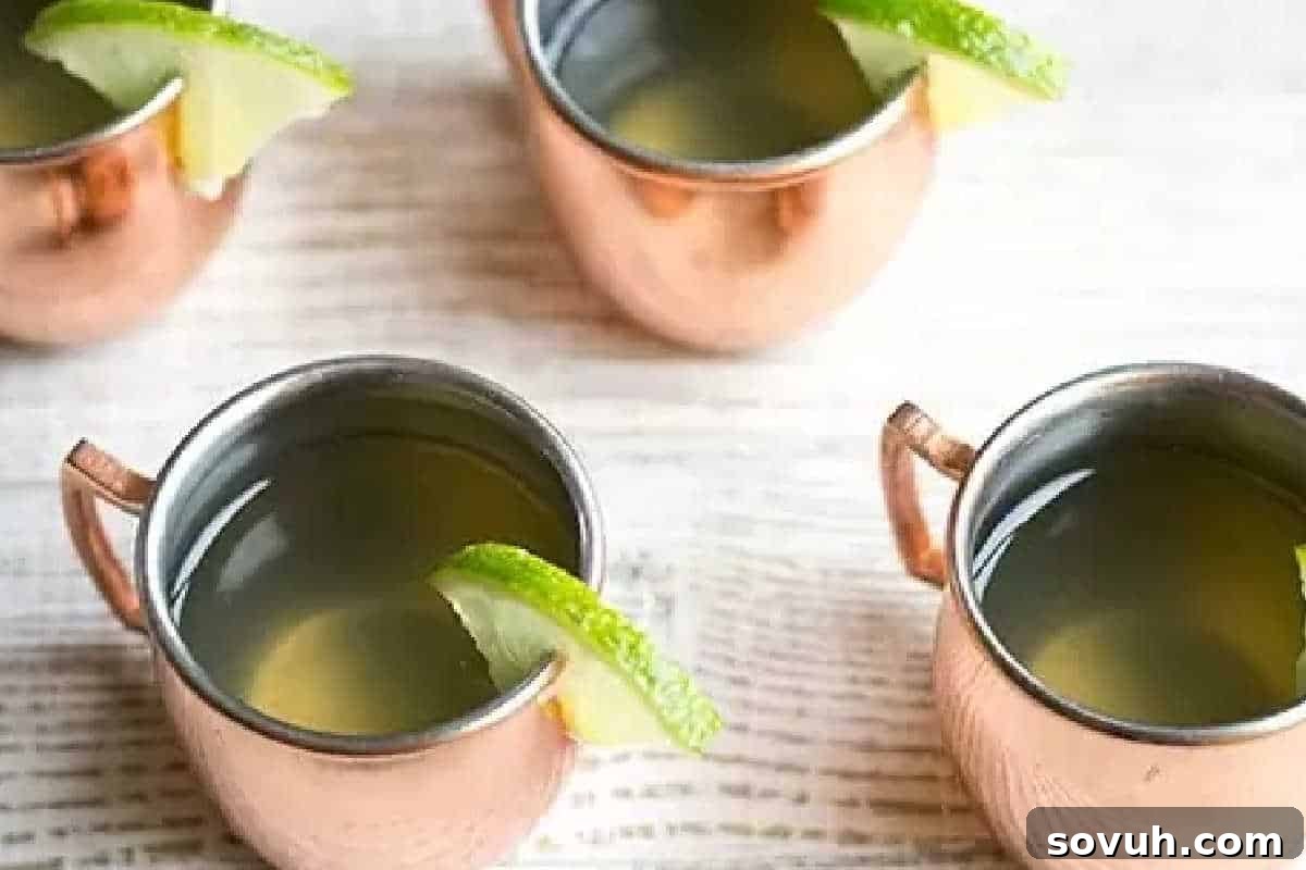 Four copper mugs filled with a light beverage, each garnished with a lime wedge, arranged on a light wooden surface.