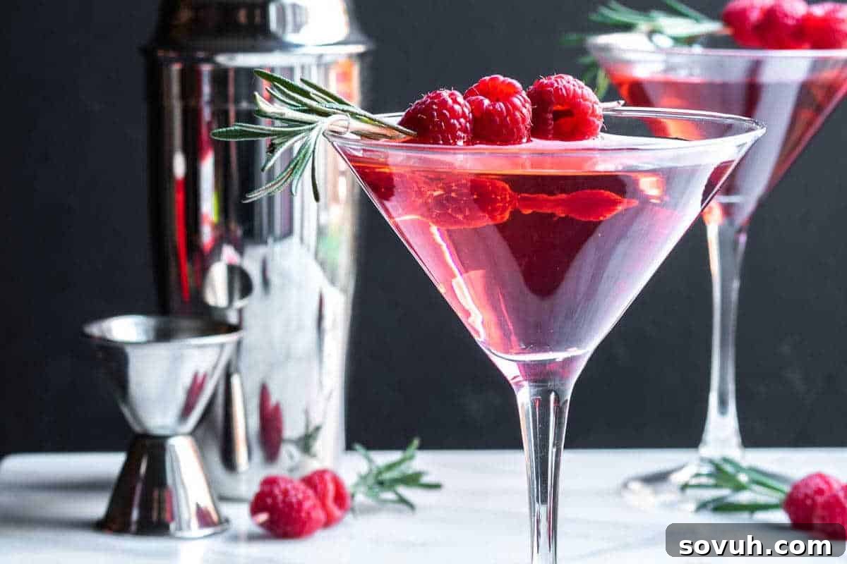 A martini glass filled with a pink cocktail, garnished with raspberries and rosemary, sits on a white surface with a cocktail shaker and jigger in the background.