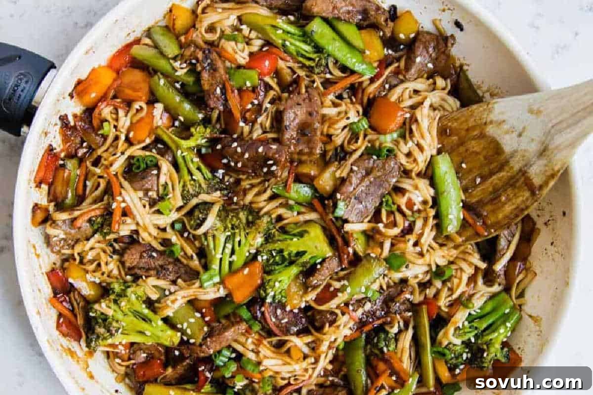 A savory skillet of stir-fried noodles with tender beef, crisp broccoli, sweet snap peas, vibrant carrots, and red bell pepper, all topped with sesame seeds and being mixed with a wooden spatula.
