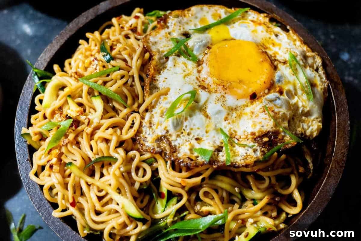 A comforting bowl of stir-fried noodles, generously topped with a perfectly cooked sunny-side-up egg and garnished with fresh sliced green onions.