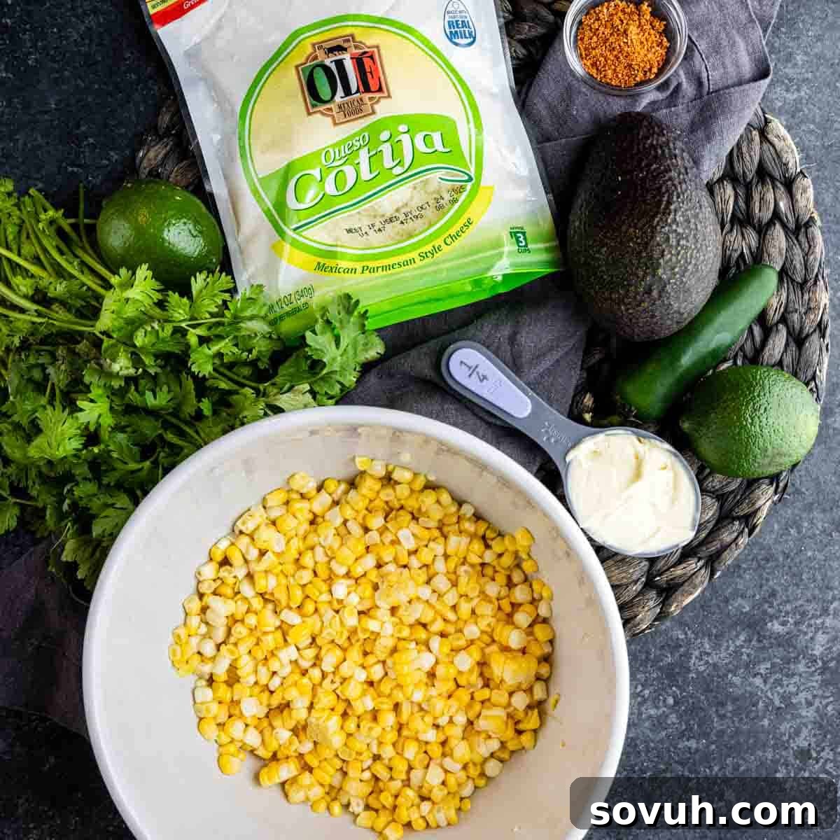 A bowl of corn kernels surrounded by cilantro, mayonnaise, avocado, lime, jalapeño, Cotija cheese, and a small bowl of seasoning on a dark surface—perfect ingredients for a flavorful Mexican street corn dip.