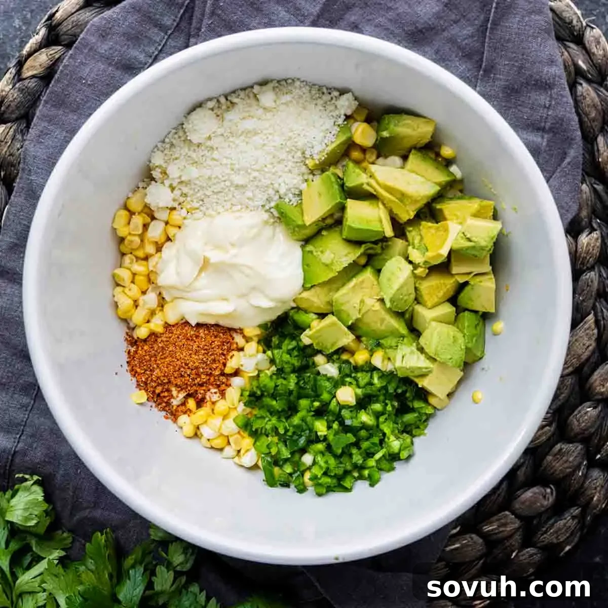 A white bowl containing diced avocado, corn, chopped jalapeños, mayonnaise, crumbled cheese, and chili powder sits on a dark napkin and woven placemat—an inviting Mexican street corn dip perfect for any gathering.