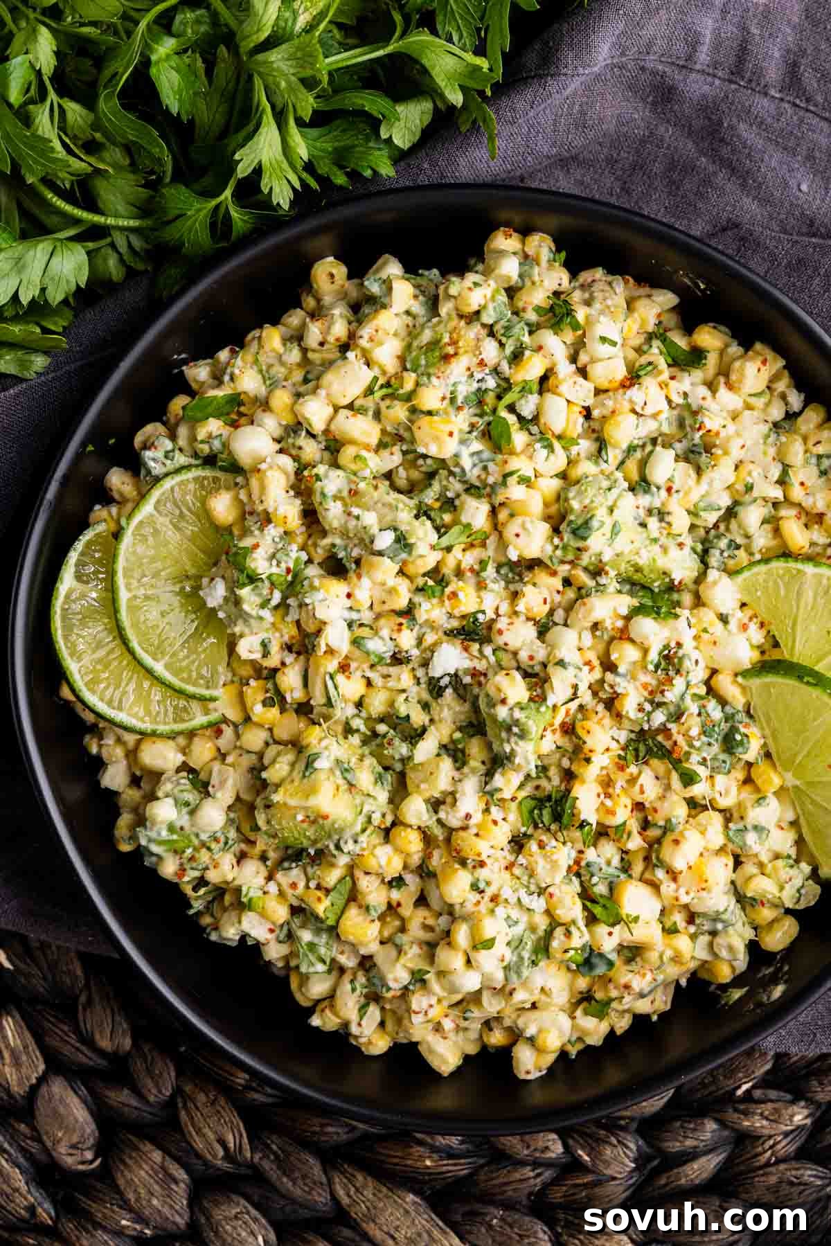 A black bowl filled with Mexican street corn salad, also known as Elote dip, garnished with lime wedges and fresh herbs, placed on a dark woven mat with parsley in the background.