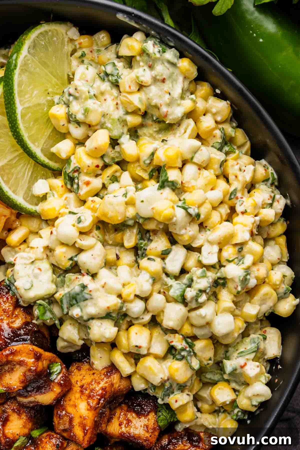 A close-up of a bowl containing creamy Mexican street corn dip with herbs and spices, garnished with lime slices, next to grilled chicken.