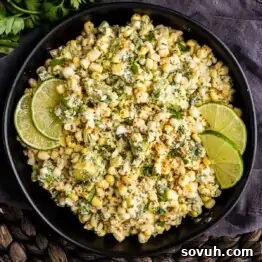 A black bowl filled with Mexican street corn salad, also known as Elote dip, garnished with lime wedges, chopped herbs, and sprinkled spices on top.