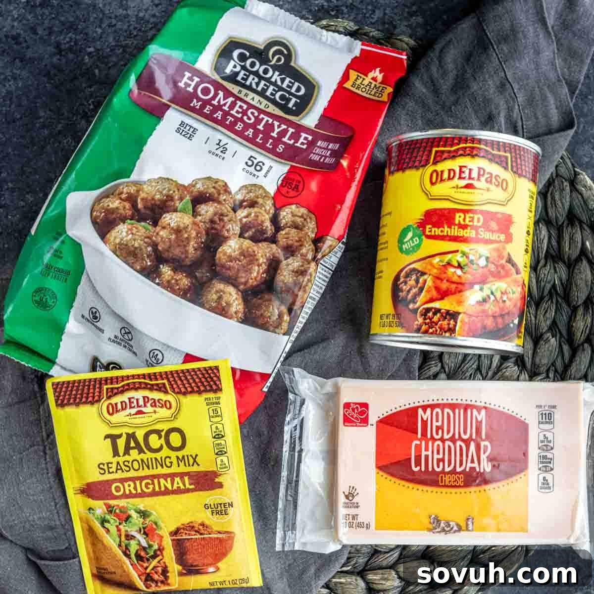 Essential ingredients for crockpot enchilada meatballs: a clear package of frozen homestyle beef meatballs, a can of vibrant red enchilada sauce, a packet of aromatic taco seasoning, and a generous pile of shredded cheddar cheese, all artfully arranged on a dark, rustic wooden surface.
