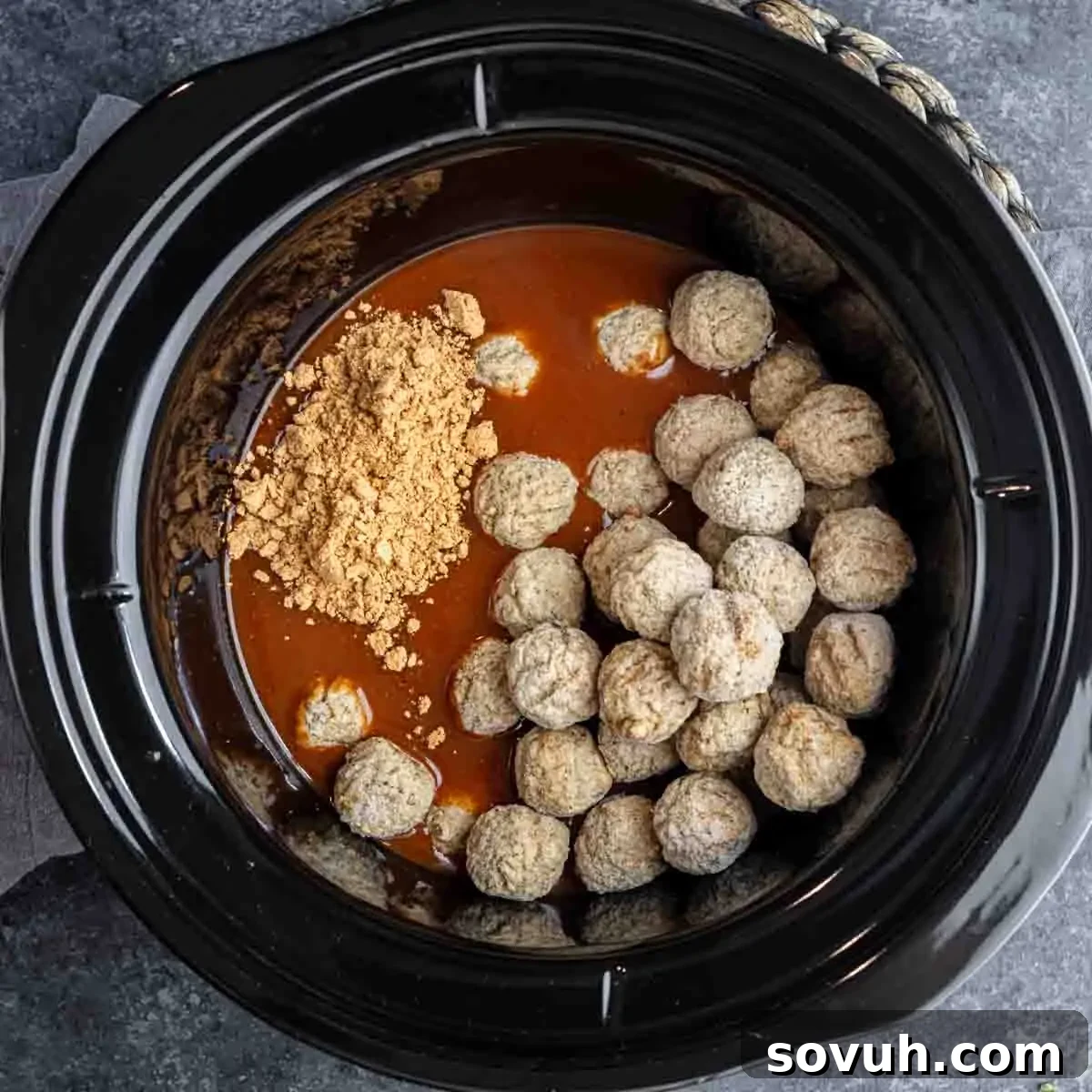 A slow cooker pot containing frozen meatballs, being covered with rich red enchilada sauce and a sprinkle of taco seasoning, illustrating the initial steps for easy slow cooker enchilada meatballs.