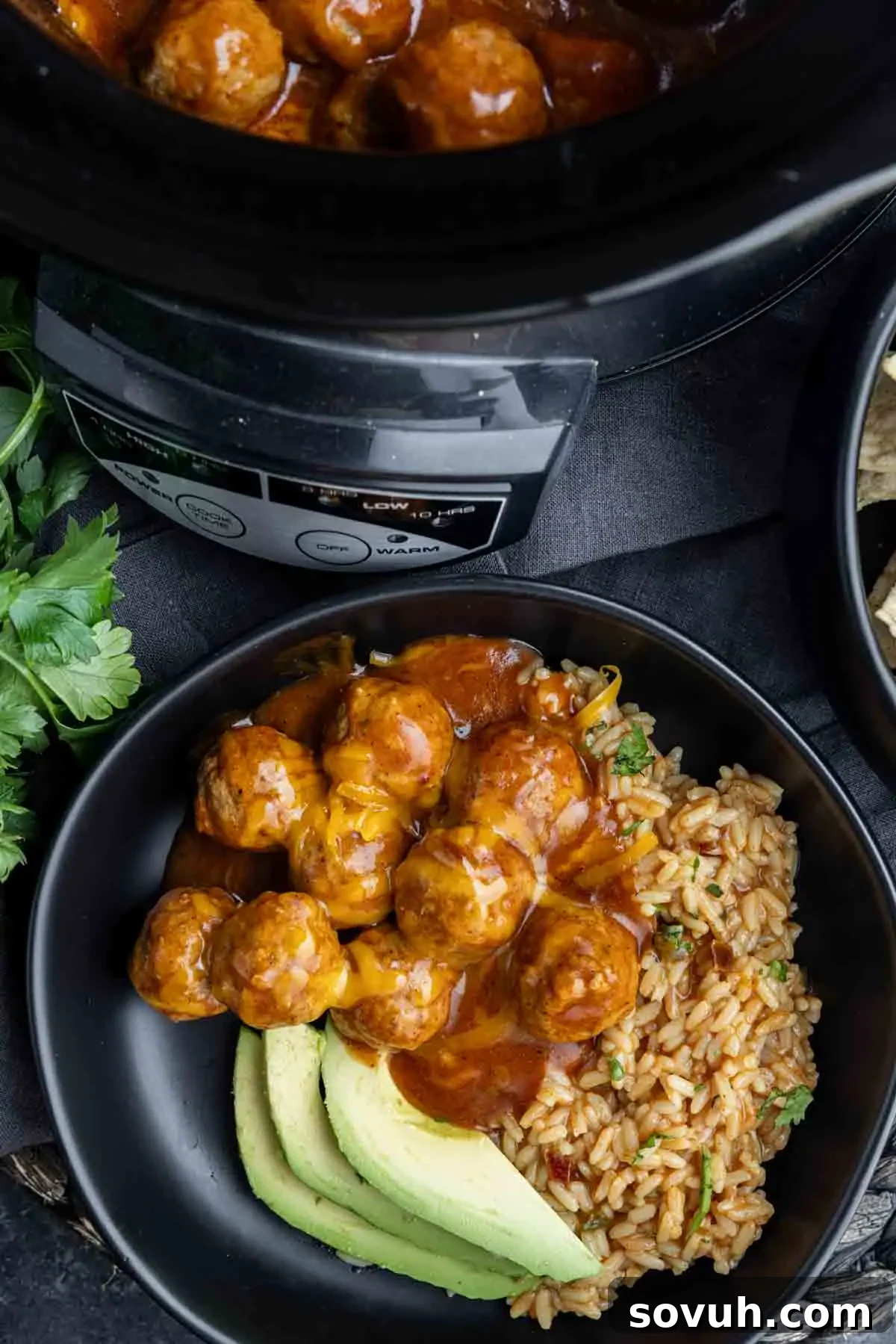 A vibrant black bowl holds saucy crockpot enchilada meatballs generously topped with shredded cheese, accompanied by seasoned Mexican rice and perfectly sliced avocado. In the background, a slow cooker peeks with more simmering meatballs.