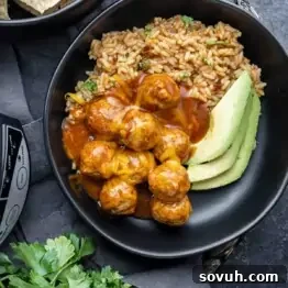 A black bowl containing meatballs in sauce with shredded cheese, sliced avocado, and a serving of seasoned rice.