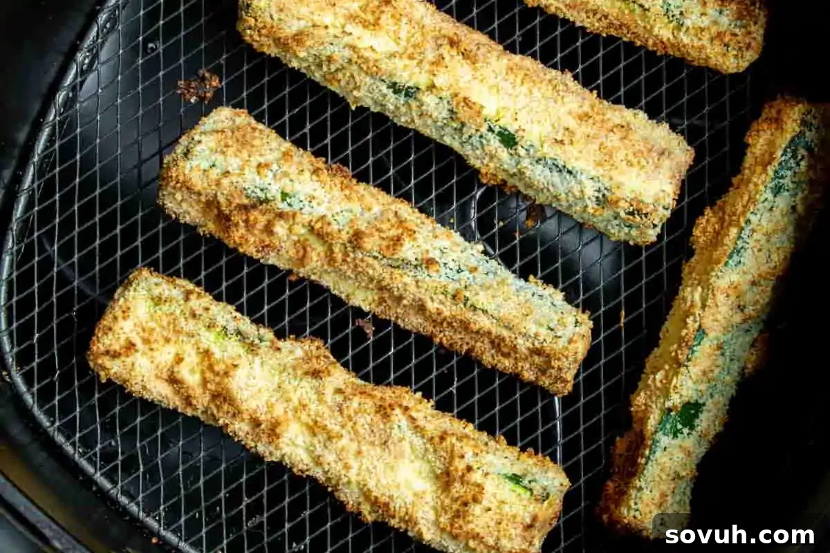 Five perfectly breaded zucchini sticks, cooked to a delectable golden-brown crisp, are neatly arranged on a black metal air fryer tray, showcasing an ideal and healthy option for your favorite zucchini recipes as a side or snack.