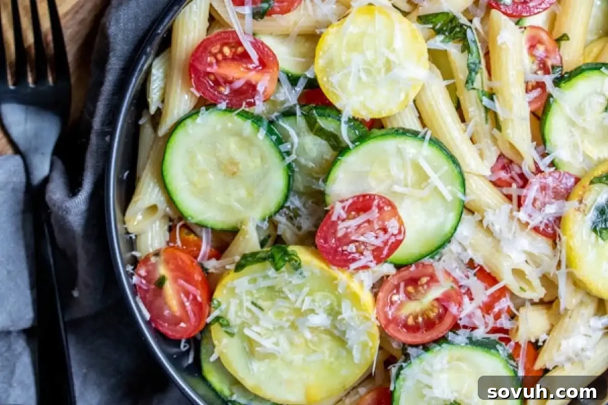 A vibrant bowl of penne pasta abundantly topped with tender sliced zucchini, bright yellow squash, juicy cherry tomatoes, and a generous sprinkle of grated cheese—perfect for those seeking new, fresh zucchini recipes. A fork and napkin are neatly arranged beside the bowl, poised for a delightful meal.