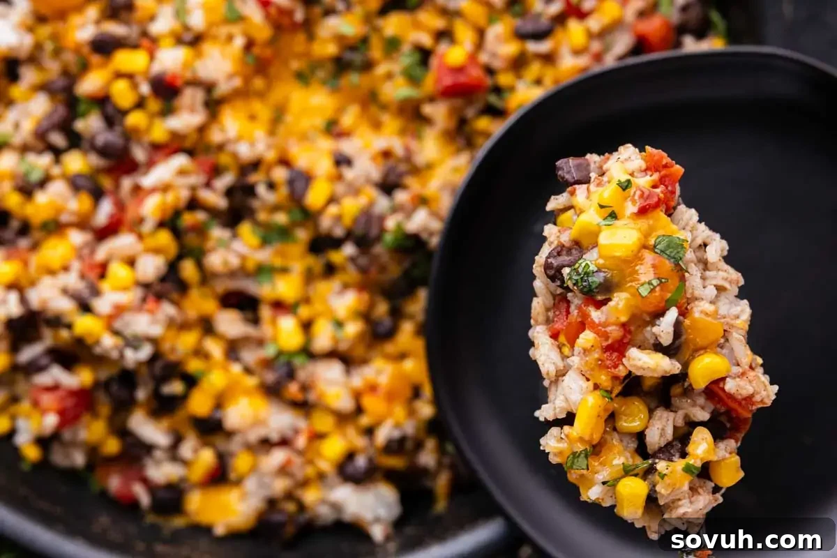 A close-up of a spoonful of rice casserole with corn, black beans, tomatoes, and melted cheese on a black plate, with the rest of the dish in the background.