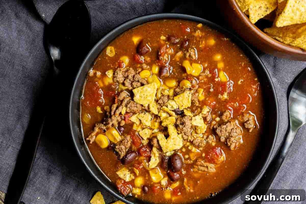 A bowl of taco soup with ground beef, beans, corn, tomatoes, and crushed tortilla chips on top, next to a spoon and a bowl of tortilla chips.