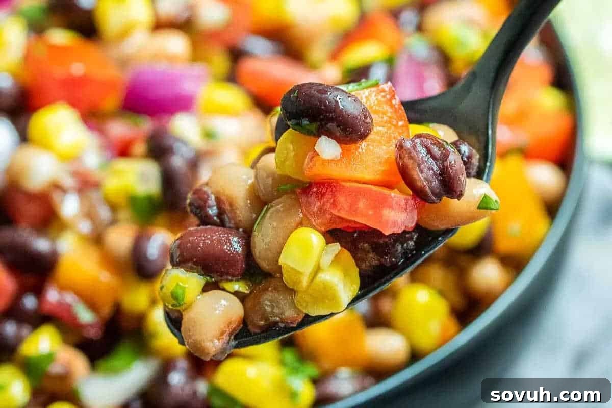 A close-up of a spoonful of colorful bean salad with black beans, black-eyed peas, corn, tomatoes, and chopped vegetables.