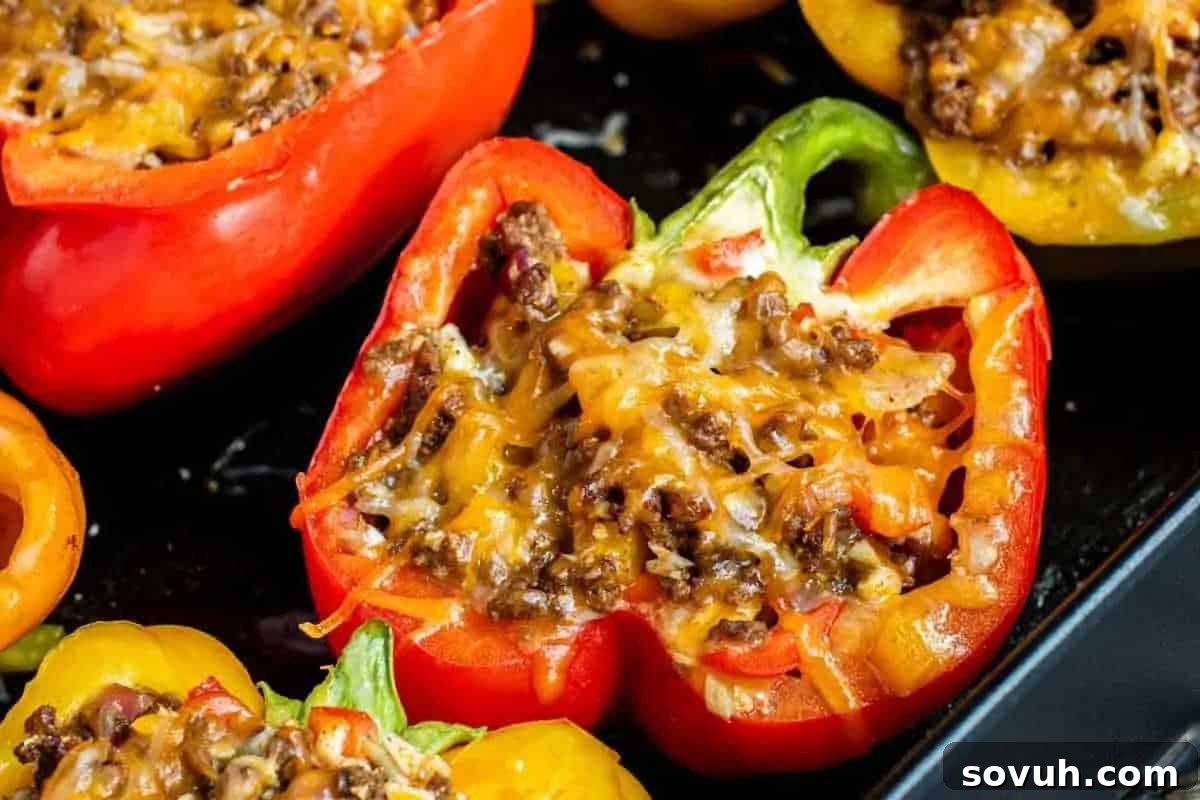 Halved bell peppers filled with a mixture of ground meat, melted cheese, and vegetables on a dark baking tray.
