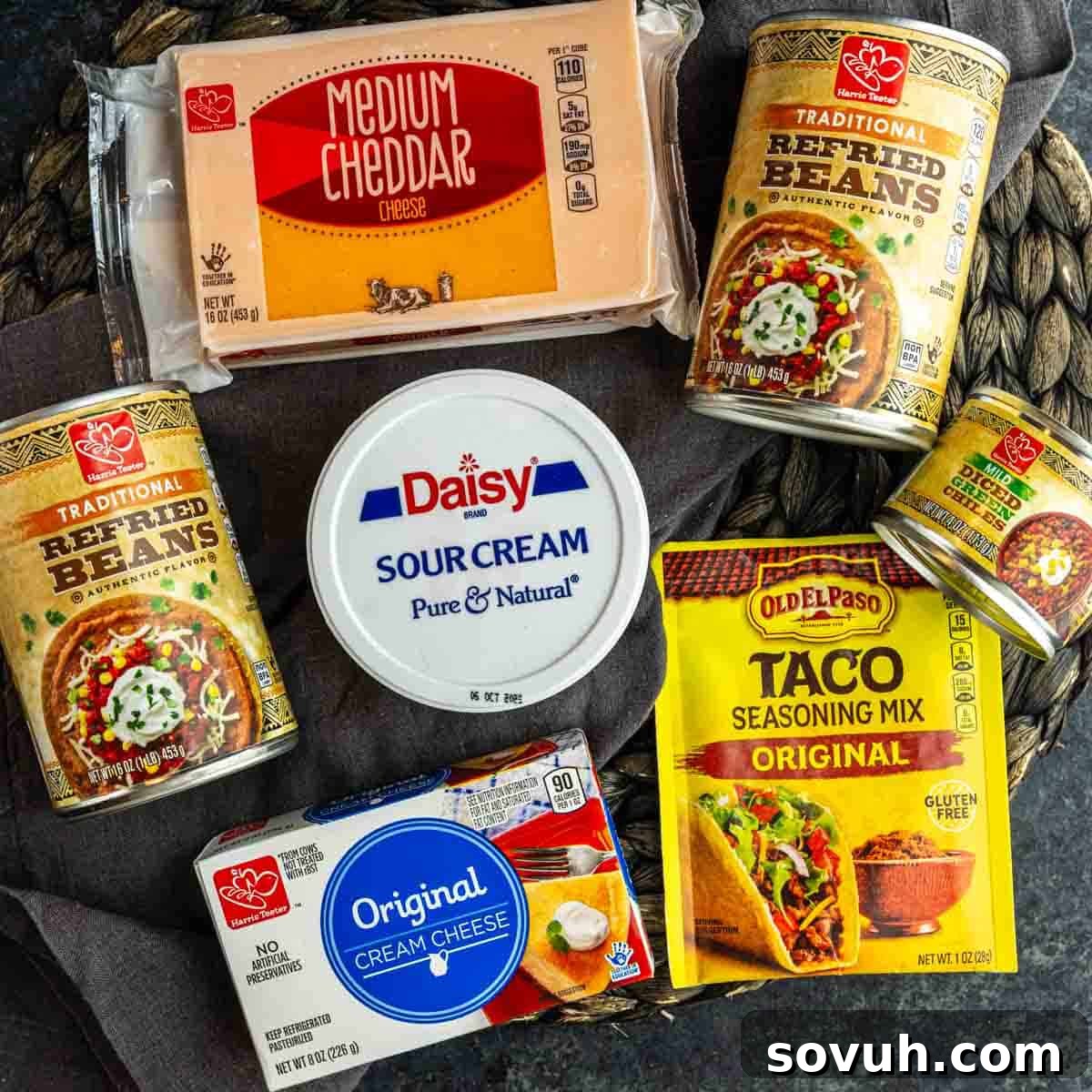Assorted taco ingredients for a cheesy bean dip or Texas trash dip, including cheddar cheese, refried beans, sour cream, cream cheese, taco seasoning mix, and diced green chiles on a dark surface.
