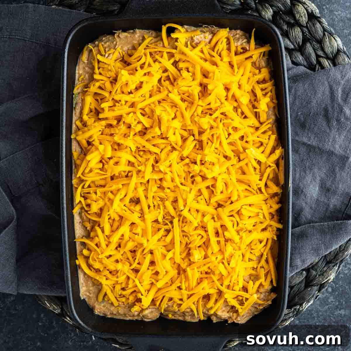 A black baking dish filled with an uncooked Texas trash dip casserole, topped with a layer of shredded cheddar cheese, placed on a dark cloth.
