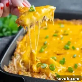 A hand lifts a cheesy slice of casserole from a baking dish, with gooey cheese stretching and chopped green onions on top—perfect for fans of Texas trash dip.