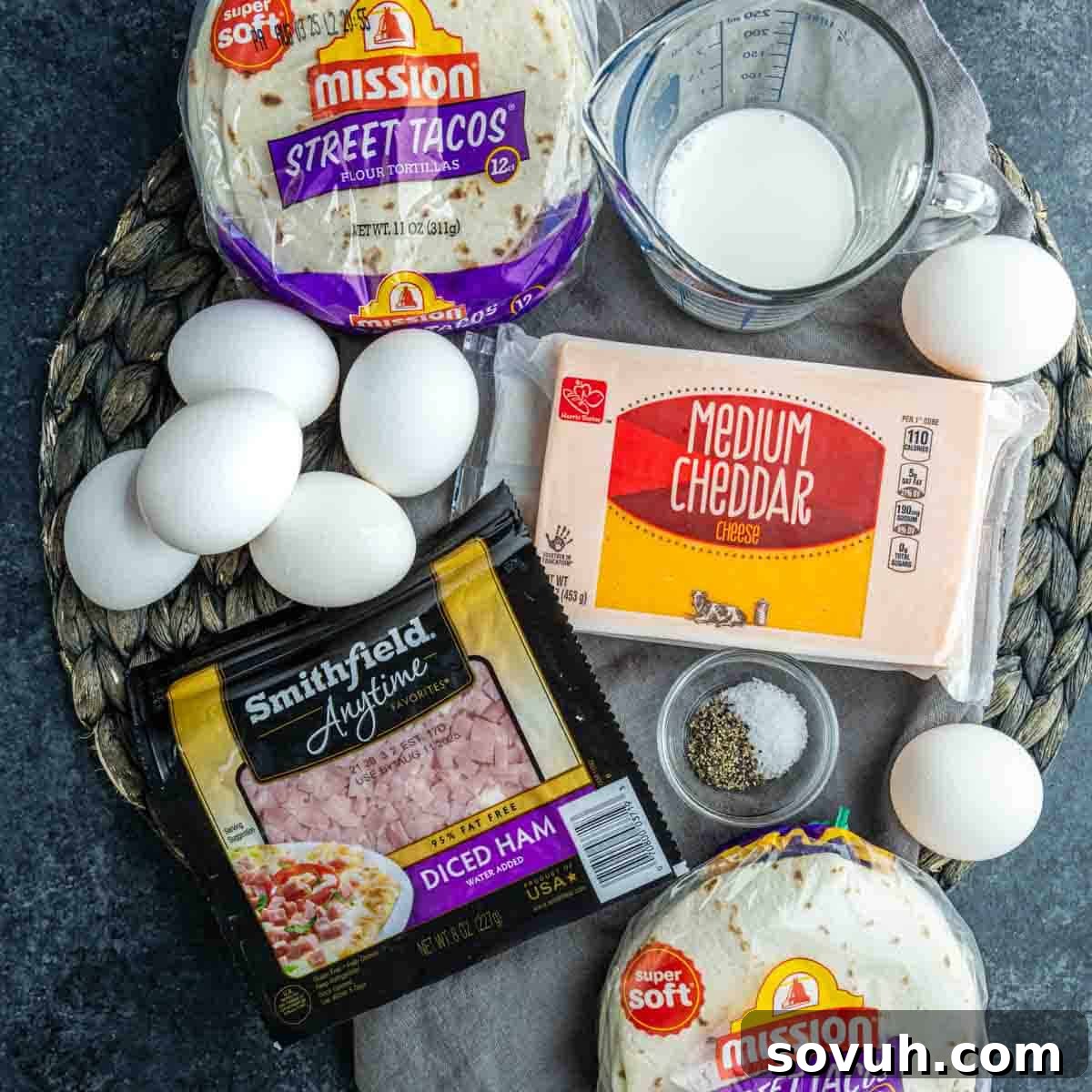 Tortillas, eggs, diced ham, cheddar cheese, milk, and seasonings are set out on a woven placemat—everything you need to whip up easy breakfast burrito cups.