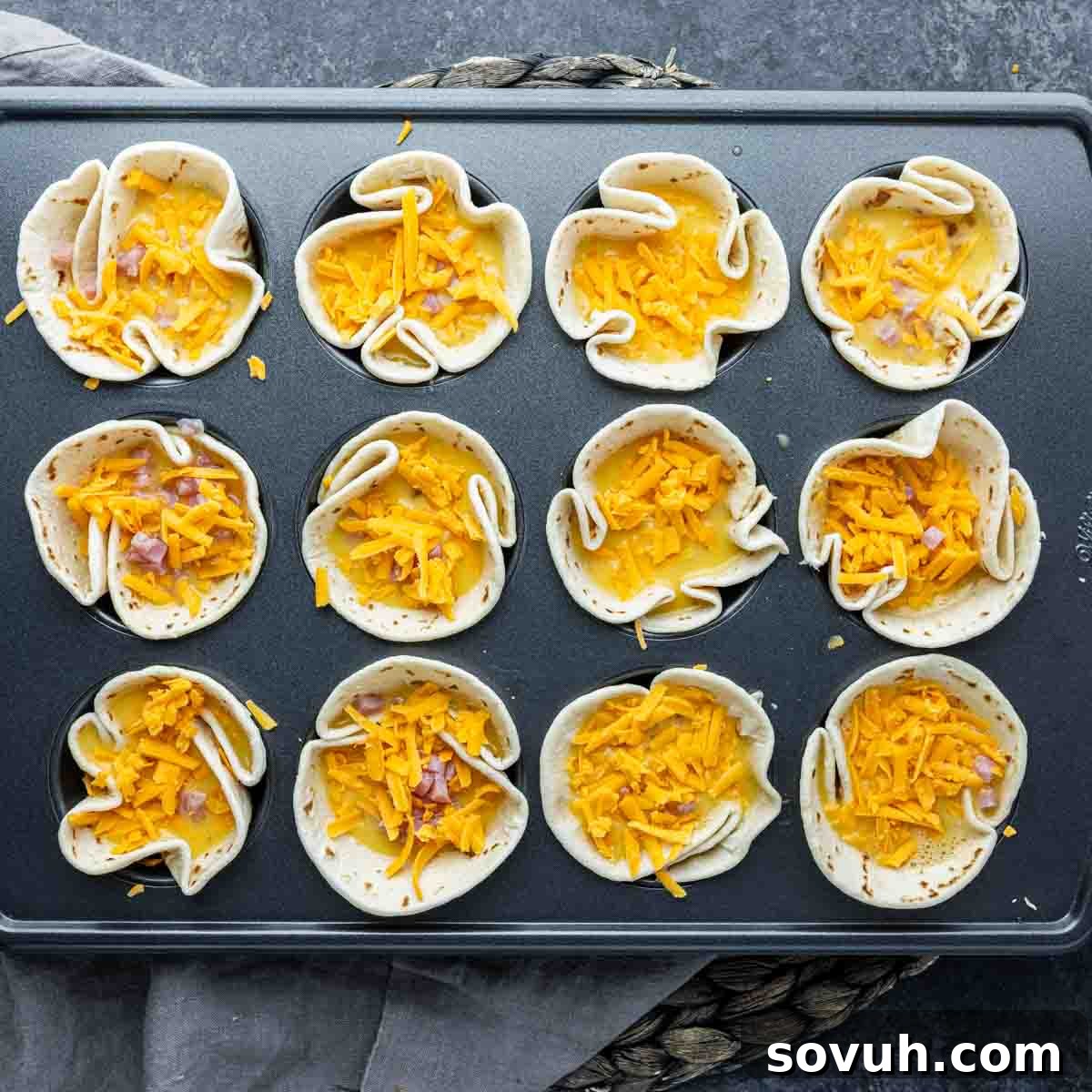 A muffin tin with twelve easy breakfast burrito cups filled with shredded cheddar cheese and diced red onion, ready for baking.