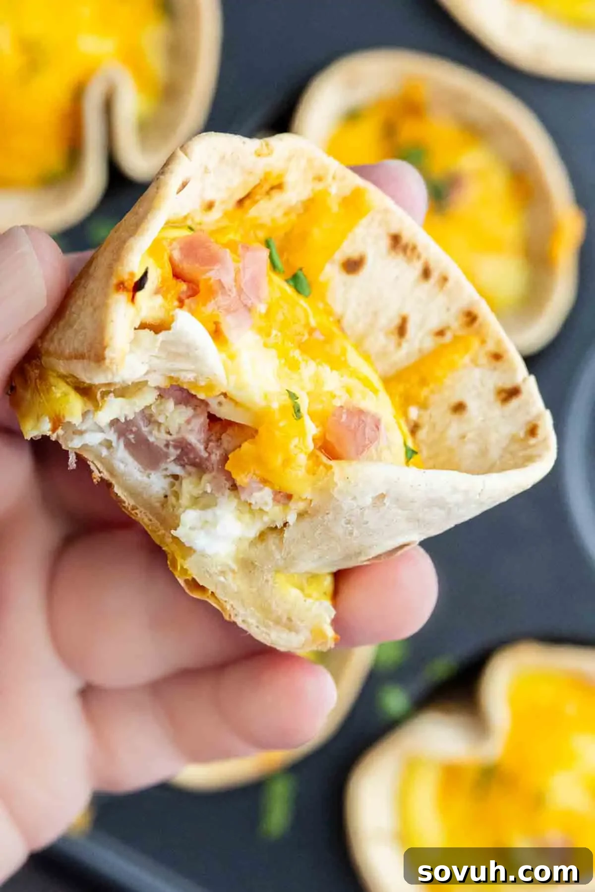 A hand holds one of these easy breakfast burrito cups—a baked tortilla filled with scrambled eggs, ham, and melted cheddar cheese—with a bite revealing its delicious layers.