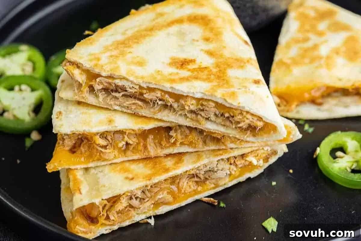 Three triangular chicken and cheese quesadillas stacked on a black plate, with sliced jalapeños on the side.