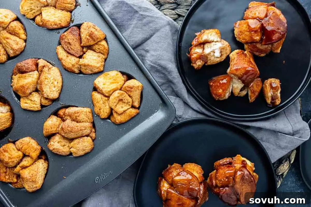 A muffin tin filled with monkey bread muffin cups sits next to three black plates, each holding a serving of monkey bread, on a dark table draped with a gray cloth.