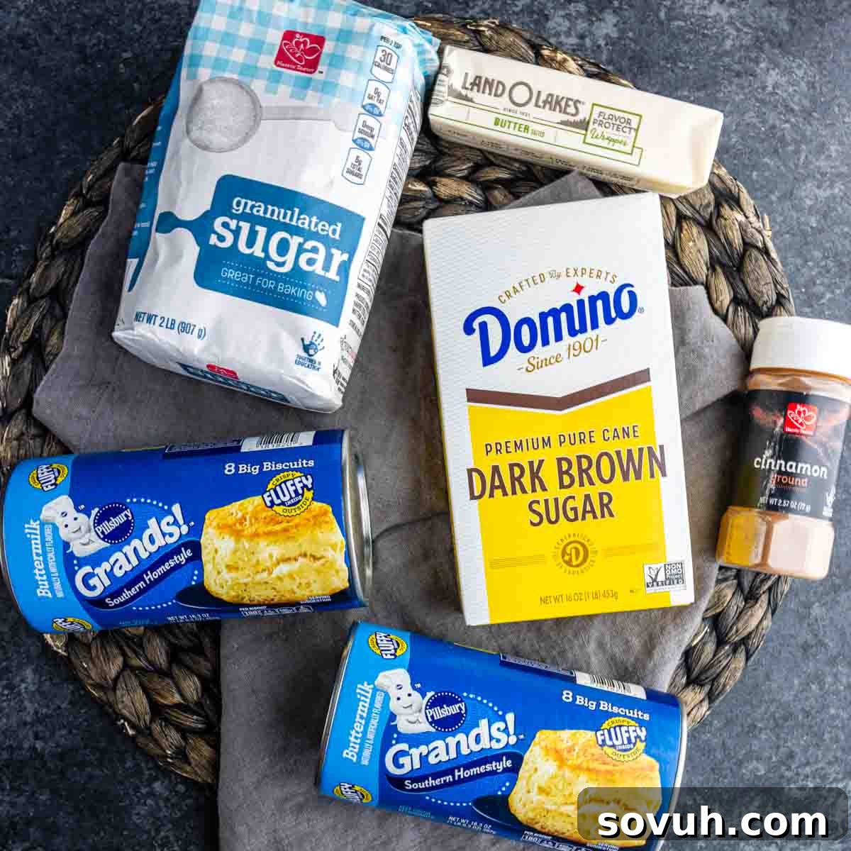 Bags of granulated and dark brown sugar, a stick of butter, ground cinnamon, and two cans of Pillsbury Grands! biscuits arranged on a woven placemat—perfect for making monkey bread muffin cups or classic monkey bread made with biscuits.
