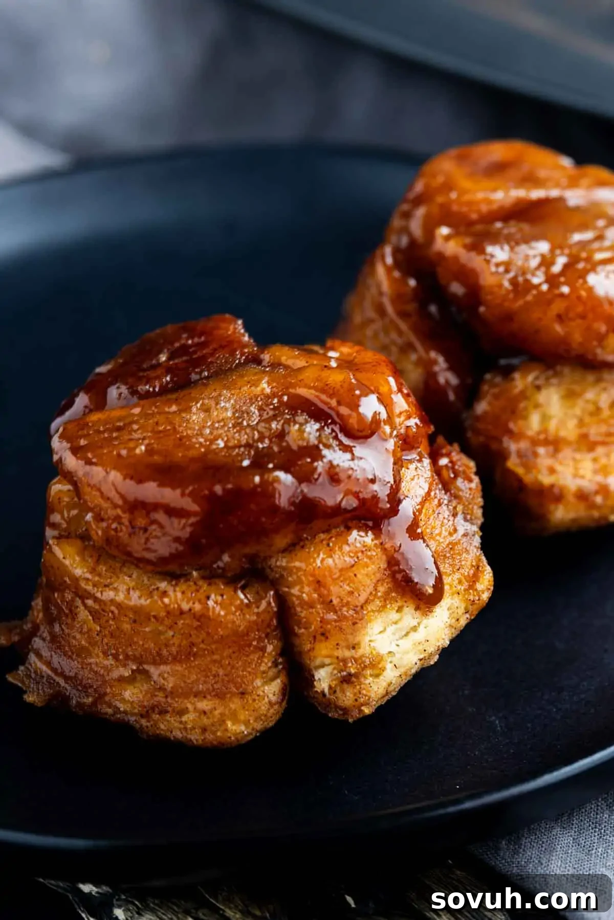 Two sticky, glazed cinnamon rolls sit on a black plate, their golden-brown, swirled layers and glossy topping reminiscent of monkey bread muffin cups.